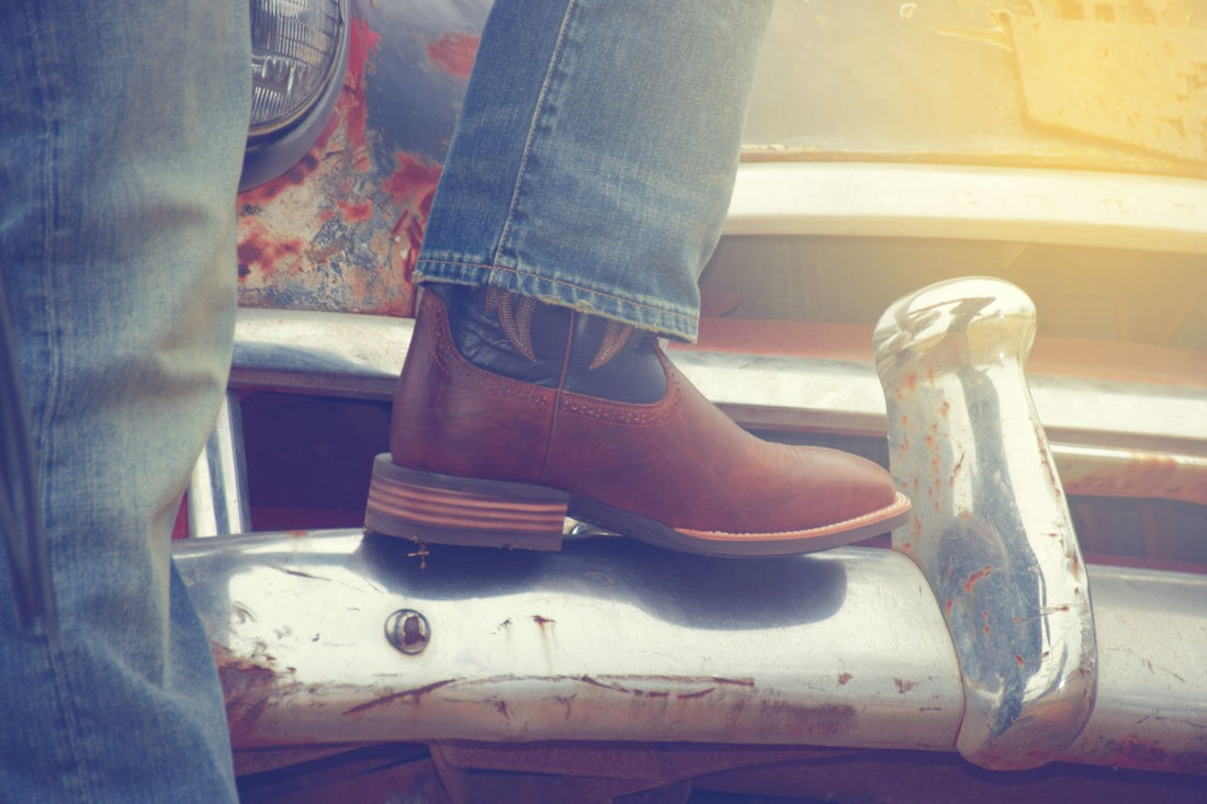 A man wearing denim jeans and brown cowboy boots rests his foot on the shiny bumper of an old rusty pickup truck.
