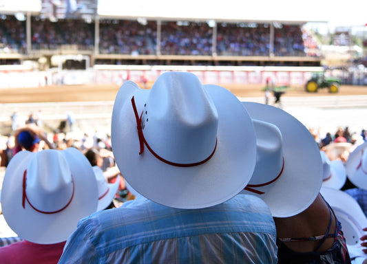 A man and woman wearing white cowboy hats watch a rodeo from the stands in a crowd with other spectators.