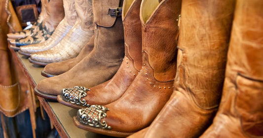 A row of high-quality leather cowboy boots, in varying heights and styles, sits on a wooden display shelf in a store.