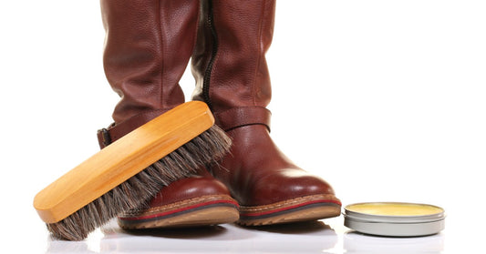 A horsehair boot brush rests on top of a pair of reddish-brown western cowboy boots next to a tin of boot wax.