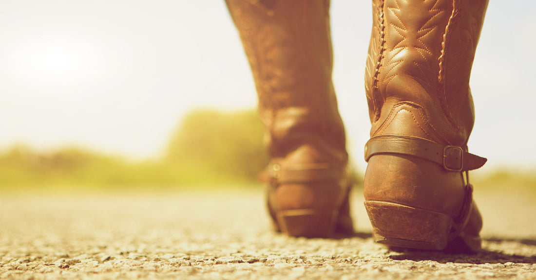 A woman wears brown leather cowboy boots with embroidery and buckle details as she walks down a gravel road.