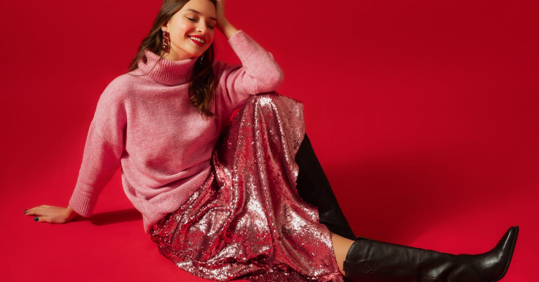 A smiling young woman wearing a pink sweater, a sequin skirt, and black cowgirl boots sits against a red background.