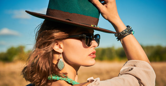 A close-up side view of a woman standing in a field holding a green cowboy hat on her head. She wears sunglasses.