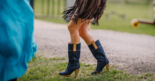 A waist-down view of a woman walking near a gravel path wearing a brown fringed dress and high-heeled cowboy boots.