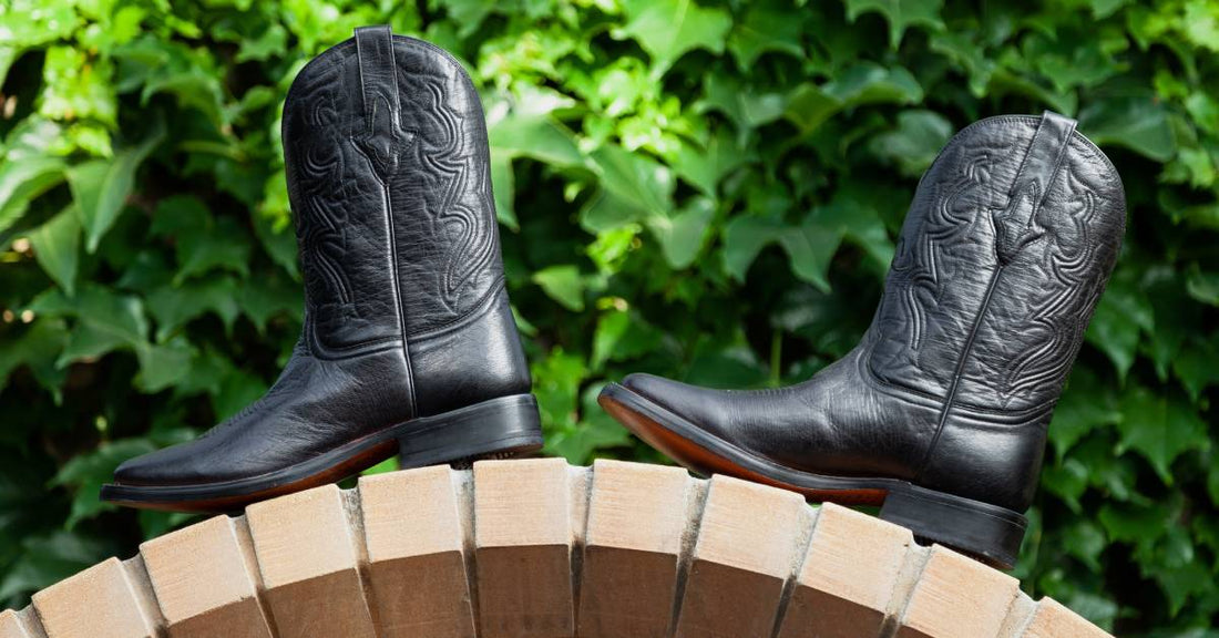 A pair of black cowboy boots sit on top of a brick archway outside in front of green ivy growing on a wall.